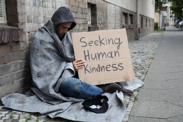 Man seated on street holding sign that reads Seeking Human Kindness